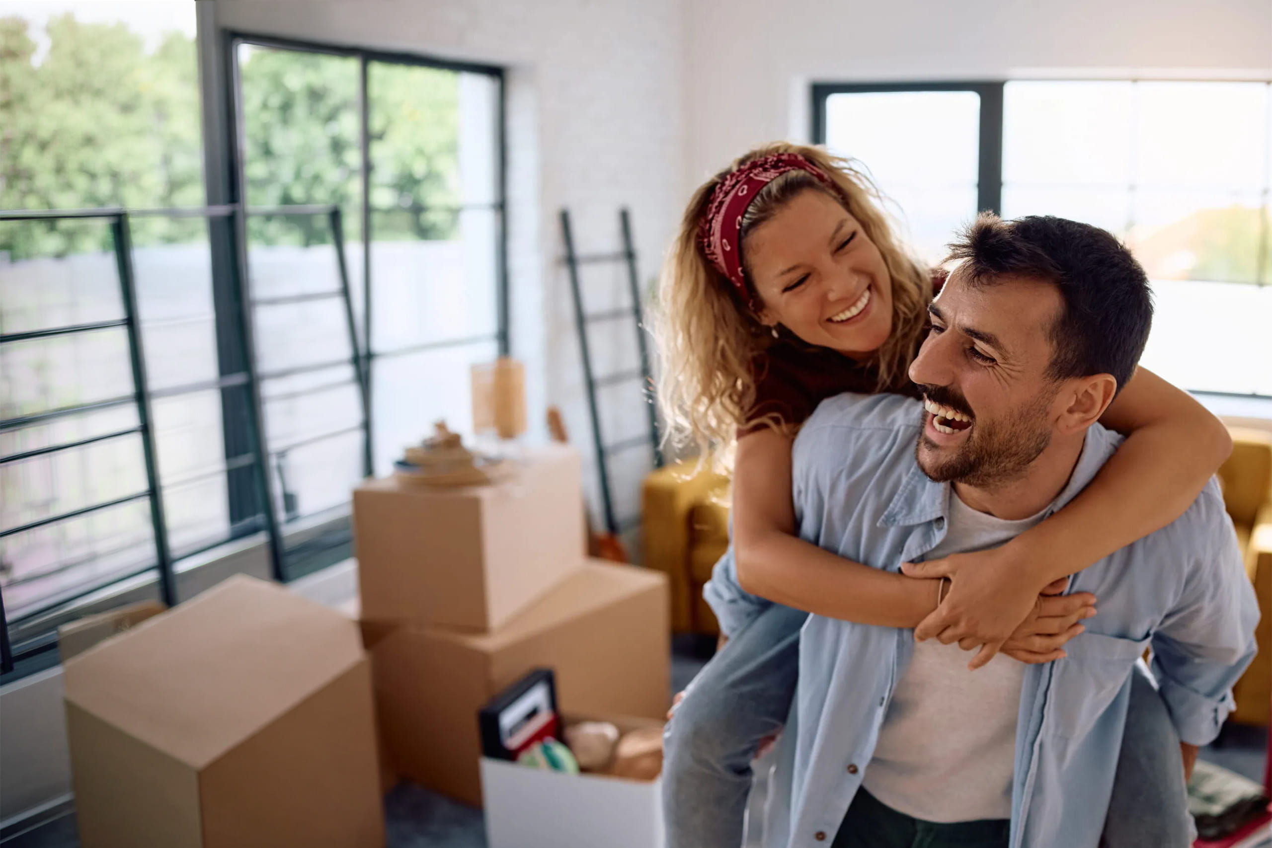 Two people in a living room surrounded by moving boxes, with one person giving the other a piggyback ride. Large windows and household items are visible in the background.