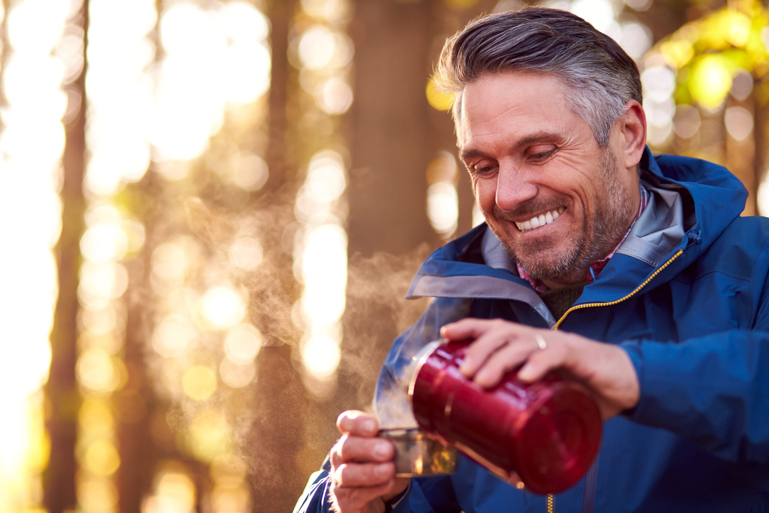 Person wearing an outdoor jacket pouring a steaming drink from a red flask into a metal cup, with trees and warm natural light in the background.