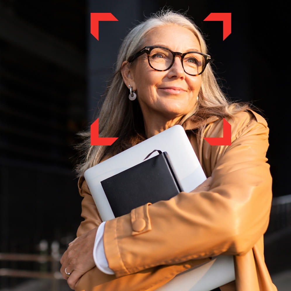 Person wearing glasses and carrying a laptop and notebook while standing outdoors beside a modern building.